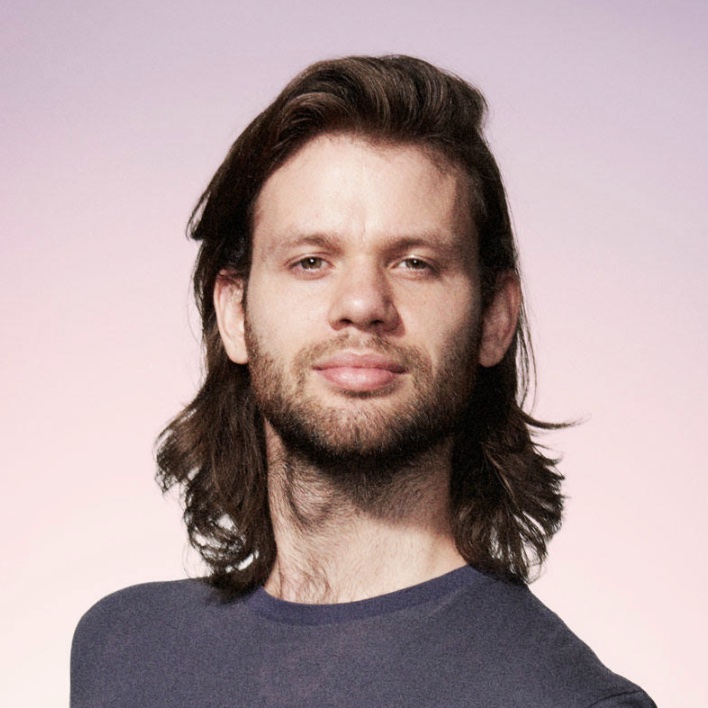 Charlie Nicholson, a softly-smiling comedian with long brown hair against a pink gradient backdrop
