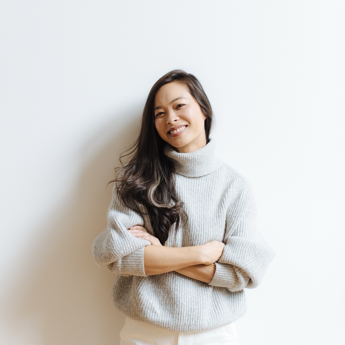 Photographer Jennifer Young in a soft grey turtleneck, arms crossed, smiling against a light studio wall